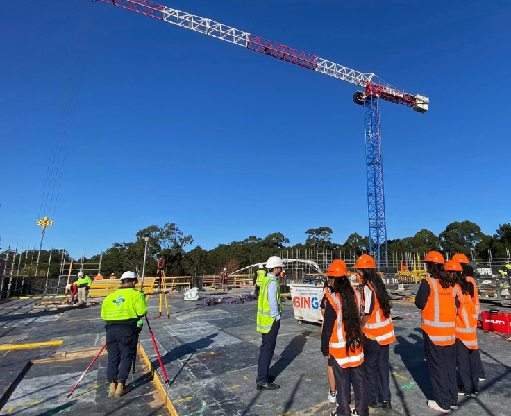 Students wearing PPE standing on worksite with workmen under crane